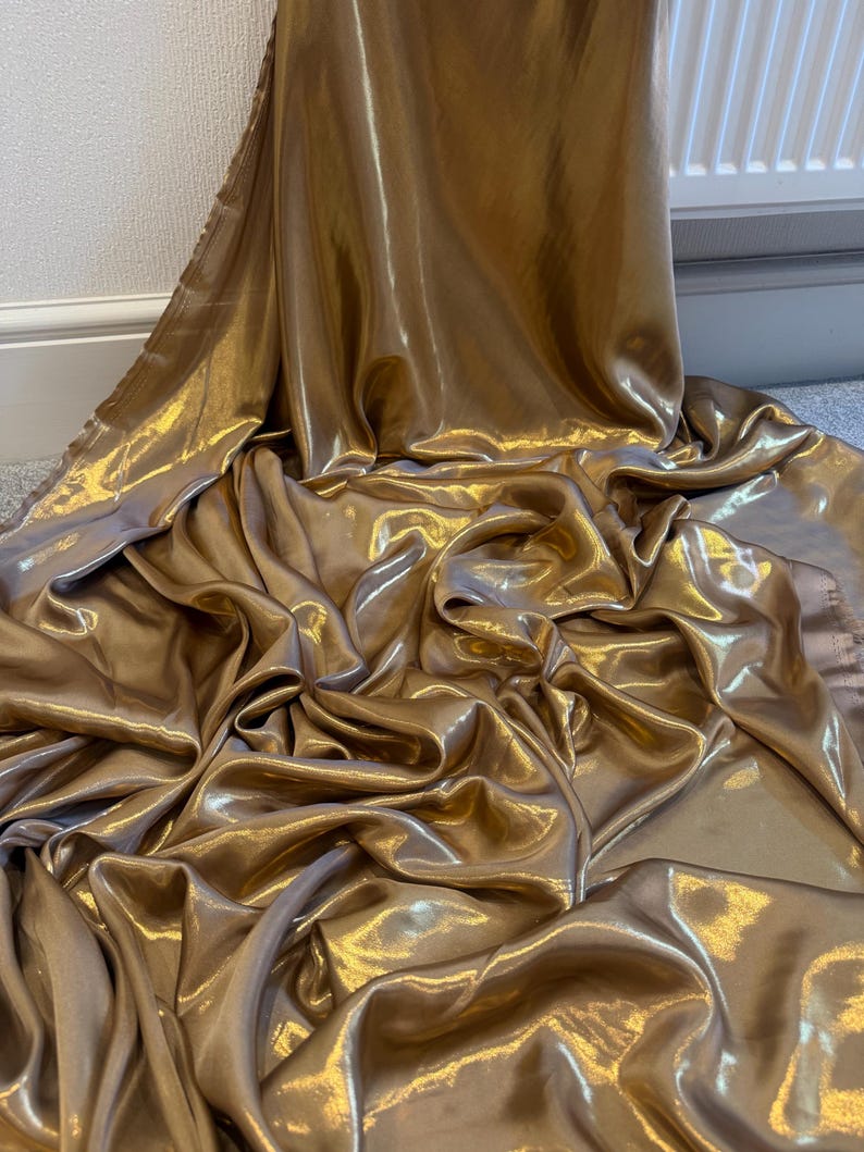 Op de afbeelding: Een luxueuze, goudkleurige stof met een glanzende satijnen afwerking. Het materiaal is gedrapeerd en gevouwen, wat een gevoel van beweging en luxe cre&euml;ert. De kleur is een warm, rijk goud, en de stof lijkt uit &eacute;&eacute;n stuk te bestaan.