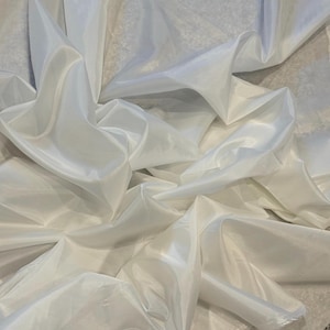 Puede incluir: Una pieza de tela blanca sólida con un acabado suave y ligeramente lustroso. El material está drapeado en pliegues suaves y fluidos, creando sombras y reflejos profundos en la superficie que enfatizan su ligereza y calidad reflectante.