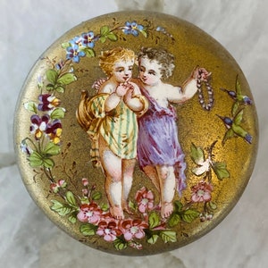 May include: A round, gold-colored trinket box with a painted scene of two children in a garden setting. The children are wearing colorful clothing and are surrounded by flowers and birds. The box is decorated with a gold-leaf finish.