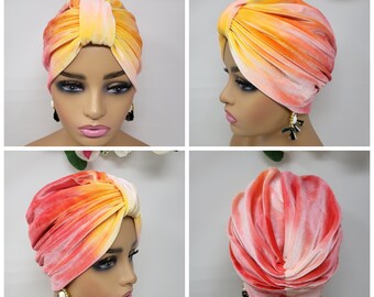 May include: A yellow and pink ombre velvet turban head wrap. The turban is styled with a knot on top and drapes down the back of the head.