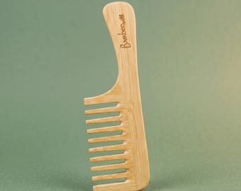Puede incluir: Un peine para el cabello de bambú de color marrón claro con un diseño de dientes anchos y un mango curvo. La palabra "bamboo" está grabada verticalmente en una fuente cursiva en la parte superior del mango, con una textura de veta natural.