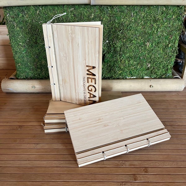 May include: A stack of natural bamboo journals with a white string closure. The top journal has the name "Megan" and the text "Thoughts & Dreams" engraved on the cover.