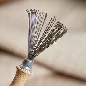 May include: A metal needle tool with a wooden handle. The tool has many thin, silver needles fanning out from a metal base. The wooden handle is light brown. The tool is likely used for cleaning.