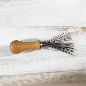 May include: A vintage brush with a wooden handle and metal bristles. The handle is light brown and rounded, with a metal ferrule connecting it to the brush head. The bristles are thin, silver-coloured wires.