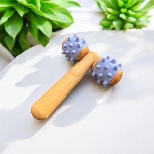 May include: A wooden massage roller with two lavender-coloured spiked heads. The roller is made of natural bamboo and has a smooth, polished finish.