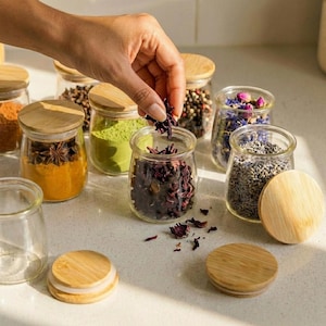 May include: A collection of small, clear glass jars with round bamboo lids, each filled with various dried herbs, spices, and floral botanicals. The jars contain ingredients such as turmeric powder, star anise, green matcha, and dried lavender.