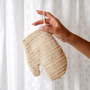 May include: A beige exfoliating bath mitt crafted from a textured, woven natural fiber material. The mitt features a thumb compartment and a small loop at the top for hanging, showcasing a coarse, open-weave construction for skin cleansing.