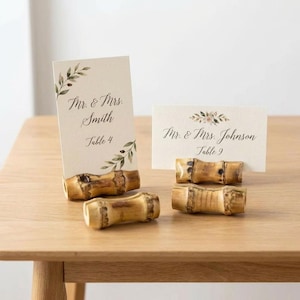 May include: Wedding place card holders made of natural bamboo. Two cards are displayed with the names "Mr. & Mrs. Smith" and "Mr. & Mrs. Johnson". The cards are decorated with floral and leaf accents. The bamboo holders are on a wooden table.