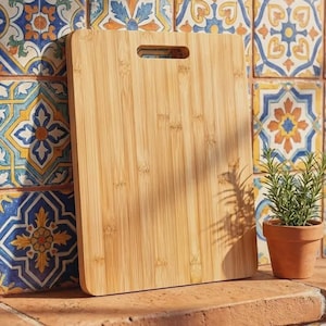 May include: A rectangular bamboo cutting board with a smooth, light brown wood grain finish and a centered handle cutout at the top. The board features vertical wood grain patterns and rounded corners, standing upright on a flat surface.