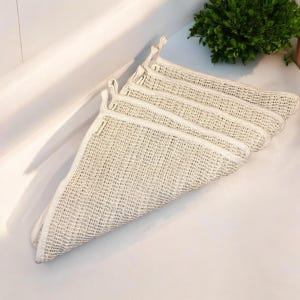May include: A set of four off-white, rectangular, mesh-textured cleaning cloths stacked in a triangular fold. Each cloth features a woven grid pattern and a small, light-colored fabric tag attached to the side edge with visible text.