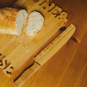 Peut inclure: Une planche à découper en bois avec le mot "INES" découpé. Un pain est partiellement tranché, avec deux tranches visibles. Un couteau à pain en bois avec un bord dentelé repose sur la planche.