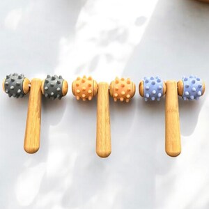 May include: Three wooden face rollers with different coloured silicone heads. The rollers are grey, orange, and purple.