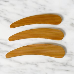 May include: Three curved, elongated wooden tools with a smooth, light brown finish and visible horizontal grain patterns. Each tool features a tapered, rounded end and a wider, slightly curved body, showcasing a natural wood texture throughout.
