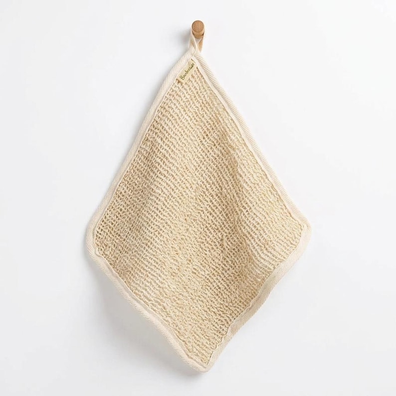 May include: A square-shaped, beige, open-weave textured washcloth with a thick, reinforced border. The cloth features a small, rectangular fabric tag with the text "Bambaw" attached to the top edge, hanging from a small wooden hook.