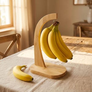 May include: A wooden banana holder with a curved top and a flat base. A bunch of yellow bananas hangs from the hook, and a single banana rests on the table. The holder is made of light-colored wood.