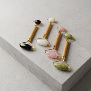 May include: Four facial rollers with bamboo handles and metal frames. One roller has black stone ends, one has white stone ends, one has pink quartz stone ends, and one has green stone ends. The metal frames vary in color between gold and rose gold.