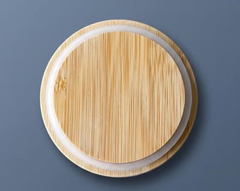 May include: A circular bamboo lid featuring a natural light wood grain pattern with vertical lines. The lid includes a white silicone gasket ring around the outer edge, designed to create a secure seal for a container or jar.
