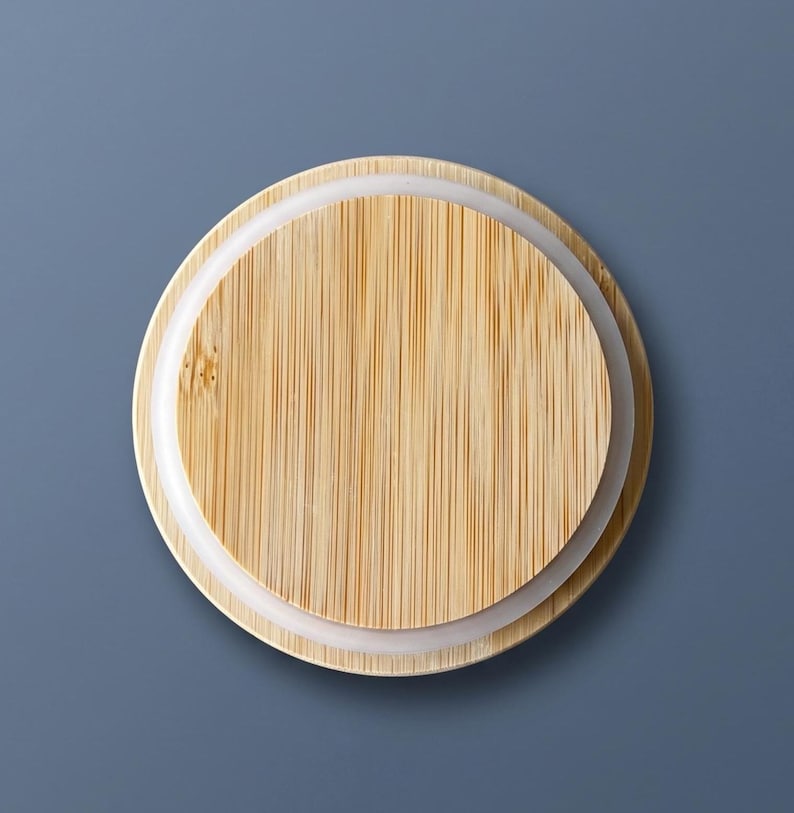 Puede incluir: Una tapa circular de bamb&uacute; con un patr&oacute;n de veta de madera clara natural y l&iacute;neas verticales. La tapa incluye un anillo de sellado de silicona blanca en el borde exterior, dise&ntilde;ado para cerrar herm&eacute;ticamente un recipiente o frasco.
