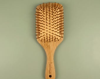 May include: A rectangular bamboo hairbrush featuring a light tan wood grain finish and numerous rounded bamboo bristles. The handle is engraved with the word bamboo in a script font and includes a small circular hole at the very bottom end.