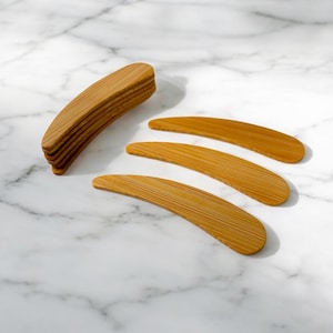 May include: Four natural bamboo spatulas, curved and slightly rounded, arranged in a fan shape on a white marble surface. The spatulas are made of light brown wood.