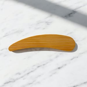 May include: A curved, flat wooden tool with a natural light brown finish and visible horizontal grain patterns. The object features a smooth, elongated shape with rounded edges, tapering slightly toward one end, showcasing a simple design.