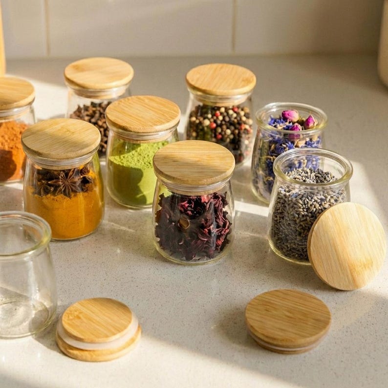 May include: A collection of clear glass jars with round bamboo lids, filled with various colorful spices, dried herbs, and floral botanicals. The jars are arranged in a group, with some lids placed separately on the surface, showcasing the contents.