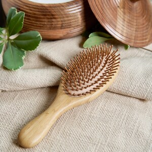 May include: A wooden hairbrush with brown bristles. The brush has an oval head and a long, curved handle. The brush is on a beige linen cloth, with a wooden bowl and leaves in the background. The brush is made of light-colored wood.