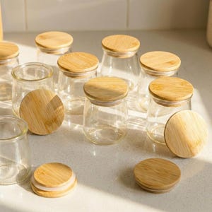 May include: A collection of small, clear glass jars with round, light-colored bamboo lids. Several jars are standing upright, while some lids are placed separately on the surface. The jars have a smooth, cylindrical shape and a wide opening.