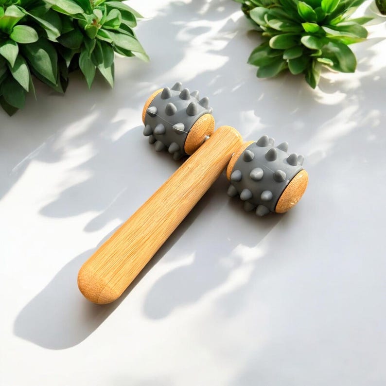 May include: A wooden massage roller with two grey spiked heads. The roller is made of natural wood and has a smooth, polished finish.