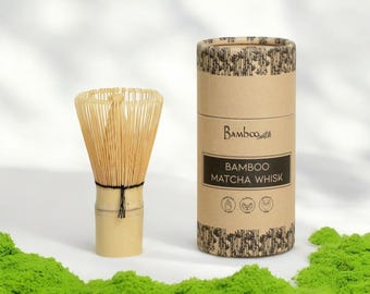 May include: A bamboo matcha whisk in a cardboard tube with the text "Bamboo Whisk" and "Bamboo Matcha Whisk". The whisk is made of natural bamboo and has a black string tied around the handle. The tube is decorated with a black and white pattern.
