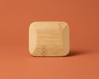 May include: A rectangular bamboo block with rounded corners and a beveled edge. The light tan wood features a visible natural grain pattern running horizontally across the surface, showcasing the organic texture and light brown color of the material.