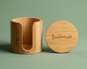 May include: A cylindrical bamboo container with a U-shaped cutout on the front side and a separate circular bamboo lid. The lid features the engraved text "Bamboowitch" in a cursive font. The natural wood grain is visible on both pieces.
