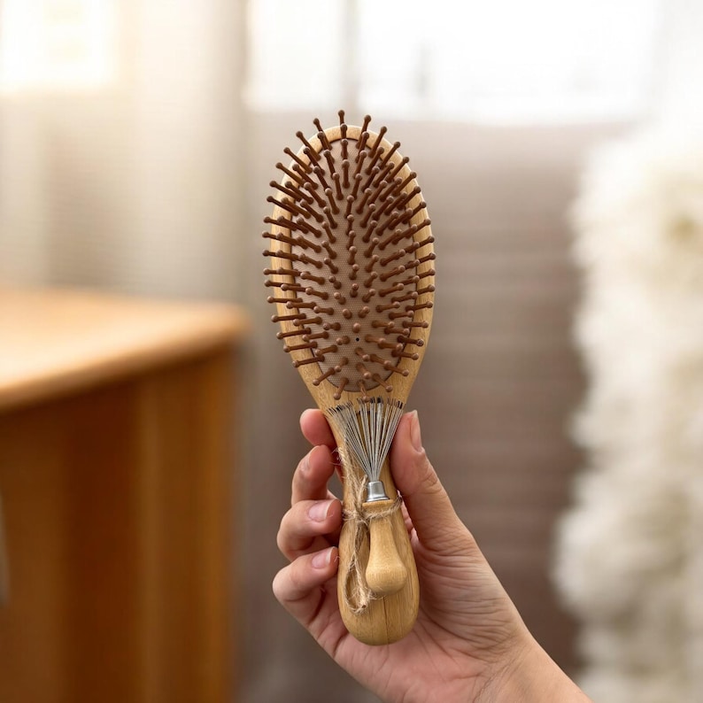 May include: Oval wooden hairbrush with brown bristles. The brush has a light wood handle and a metal and string cleaning tool attached. The brush is held in a person's hand.