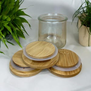 May include: A clear glass jar with a wide opening and a set of four circular bamboo lids. Each lid features a light brown wood grain pattern and a translucent silicone ring seal around the edge, designed to fit securely onto the glass container.