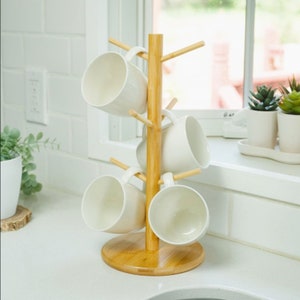 May include: A wooden mug tree with five white ceramic mugs hanging from it. The mug tree is made of natural bamboo and has a round base.