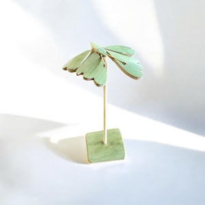 May include: A wooden decorative moth sculpture. The moth is painted in a light green color and has a square base. The wings are detailed with a scalloped edge. The moth is attached to a thin wooden dowel.