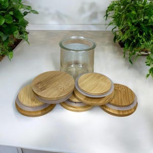 May include: A clear glass jar with a wide opening, accompanied by five circular bamboo lids. Each lid features a natural wood grain pattern and a silicone seal ring around the edge, designed to fit securely onto the top of the glass container.