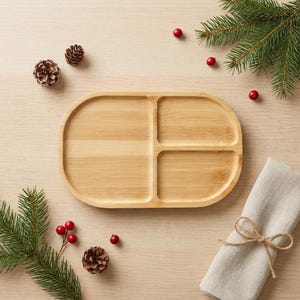 May include: A light brown, oval-shaped bamboo serving tray with three compartments. The tray is surrounded by pine needles, red berries, a pine cone, and a folded beige napkin tied with twine. Ideal for serving snacks or appetizers.