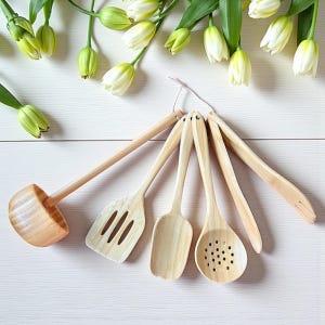 May include: A collection of six bamboo kitchen utensils, including a ladle, slotted spatula, solid spoon, slotted spoon, and two other tools. The utensils are arranged on a white wooden surface, with white and green flowers in the background.