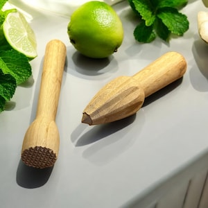 May include: Two wooden kitchen tools, a lime squeezer and a muddler, on a gray surface with a lime and mint leaves.