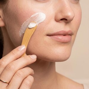 May include: A small, light brown wooden spatula with a smooth, curved handle and a flat, rounded tip, used to apply a thick, white, opaque cream onto the skin of a cheek. The wooden tool features a visible grain pattern throughout its surface.