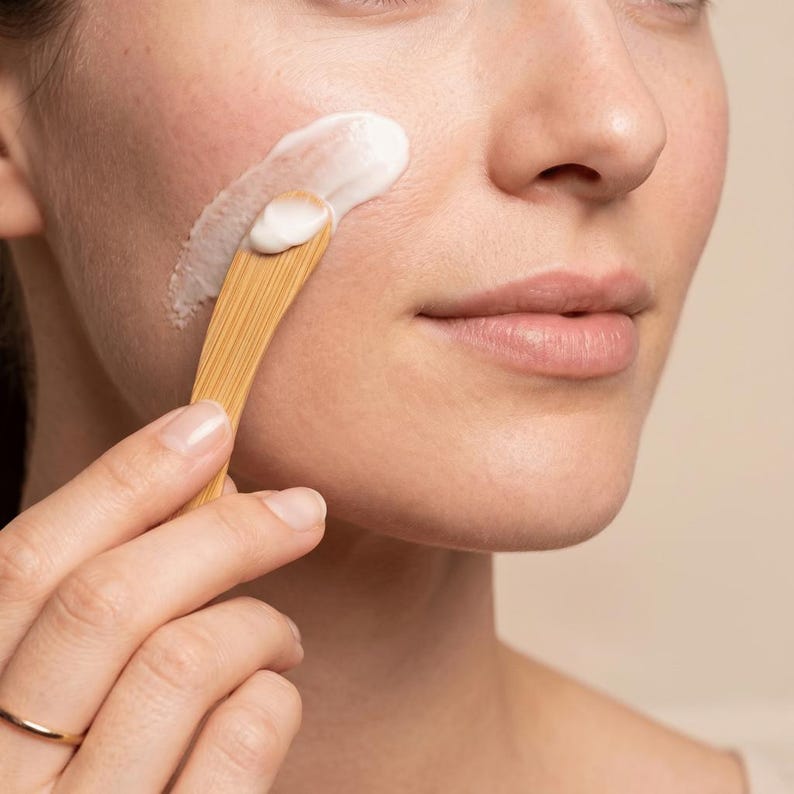 May include: A small, light brown wooden spatula with a smooth, curved handle and a flat, rounded tip, used to apply a thick, white, opaque cream onto the skin of a cheek. The wooden tool features a visible grain pattern throughout its surface.