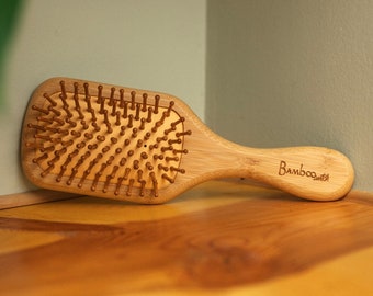 May include: A wooden hair brush with natural bristles. The brush has a rectangular shape and a brown wooden handle with the word "Bamboo" engraved on it.