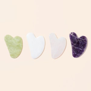 May include: Four heart-shaped gua sha tools arranged in a row. The first is light green with a mottled texture, the second is solid white, the third is pale pink, and the fourth is deep purple with white veining throughout the stone surface.
