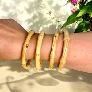 May include: Four natural bamboo bangles stacked on a wrist. The bangles are smooth and light brown in colour.