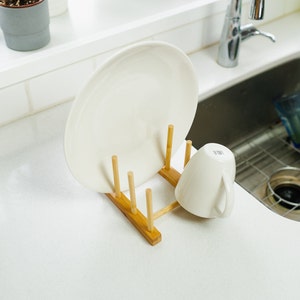 May include: A white ceramic plate and a white ceramic mug are drying on a wooden dish rack. The rack has five wooden dowels and a rectangular base.