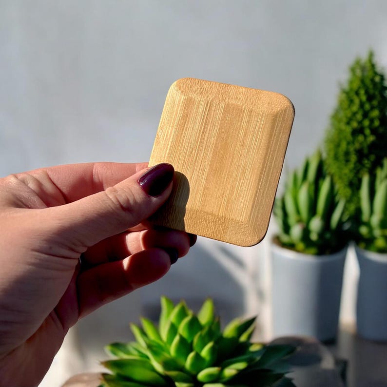 May include: A square bamboo coaster with rounded corners and a natural light brown wood grain finish. The coaster features a slightly raised border around the perimeter, showcasing the smooth texture and organic patterns of the bamboo material.