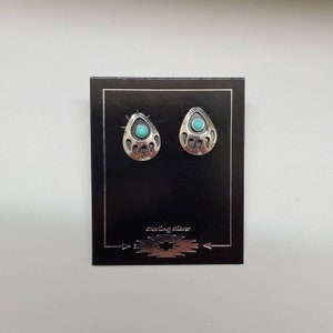 Puede incluir: Un par de pendientes de pata de oso de plata de ley con piedras turquesa. Los pendientes se muestran en una tarjeta negra con el texto "sterling silver".