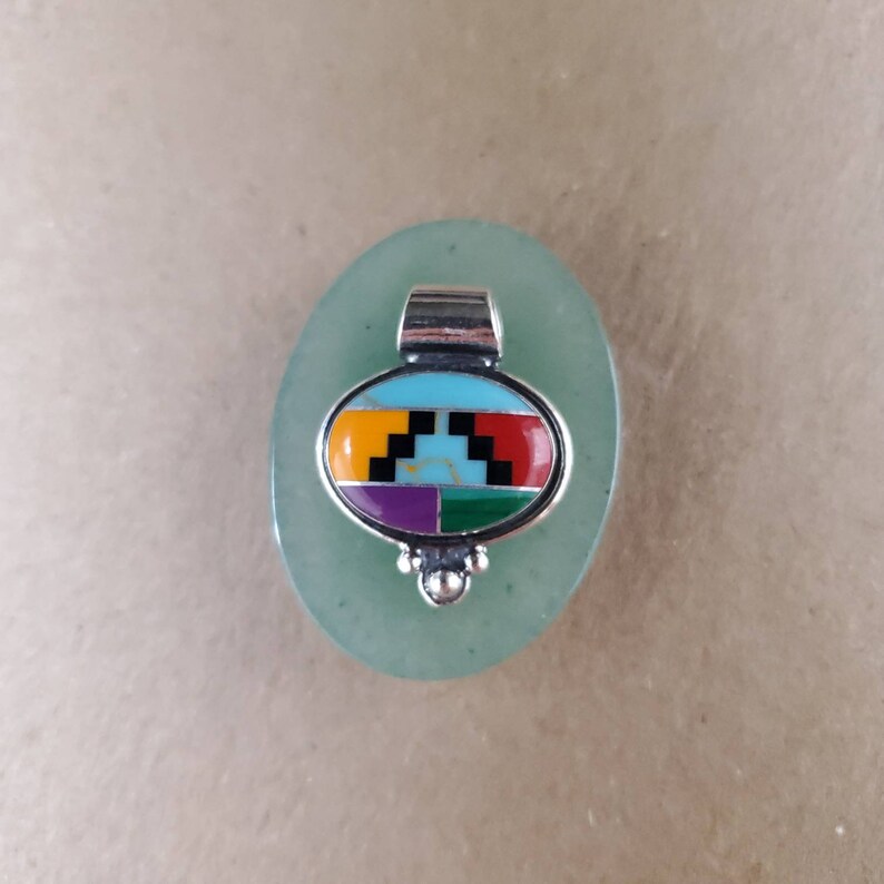 May include: A silver pendant with a turquoise, red, yellow, green, purple, and black inlay design. The pendant is oval shaped and has a bail for hanging.