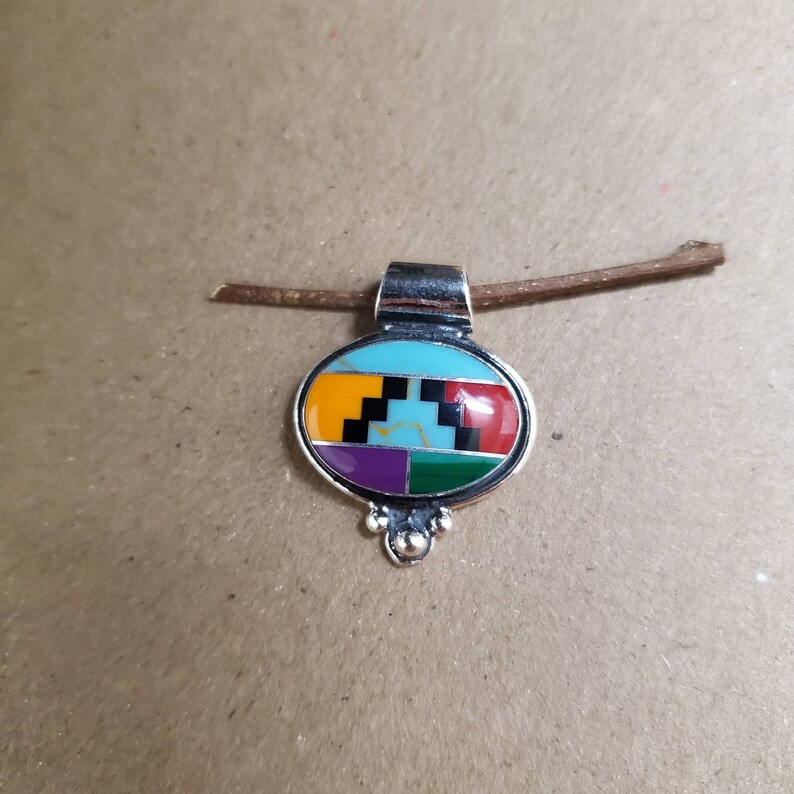 May include: A silver pendant with a colorful geometric design. The pendant is oval shaped and features turquoise, yellow, orange, red, purple, green, and black stones.